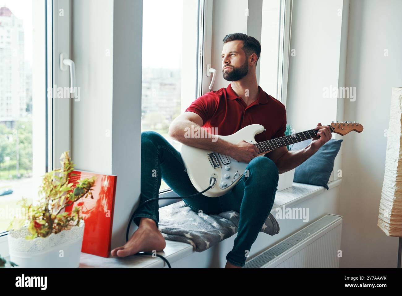 Romantic young man in casual clothing playing guitar while sitting on the window sill Stock Photo
