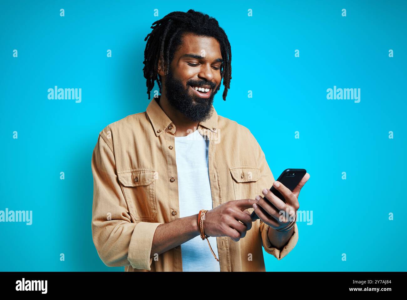 Handsome young African man in casual clothing using smart phone and smiling while standing against blue background Stock Photo