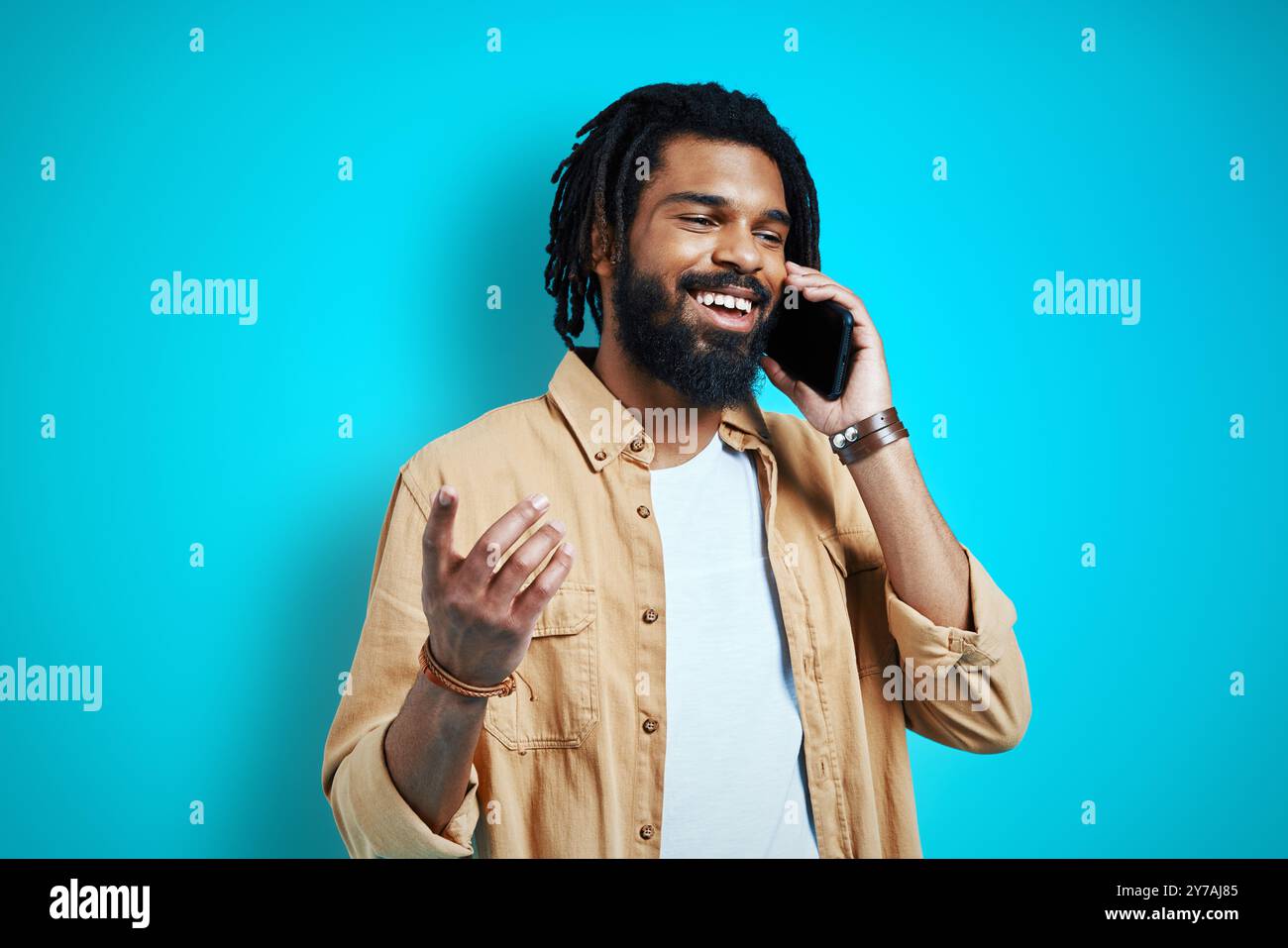 Handsome young African man in casual clothing talking on smart phone and smiling while standing against blue background Stock Photo
