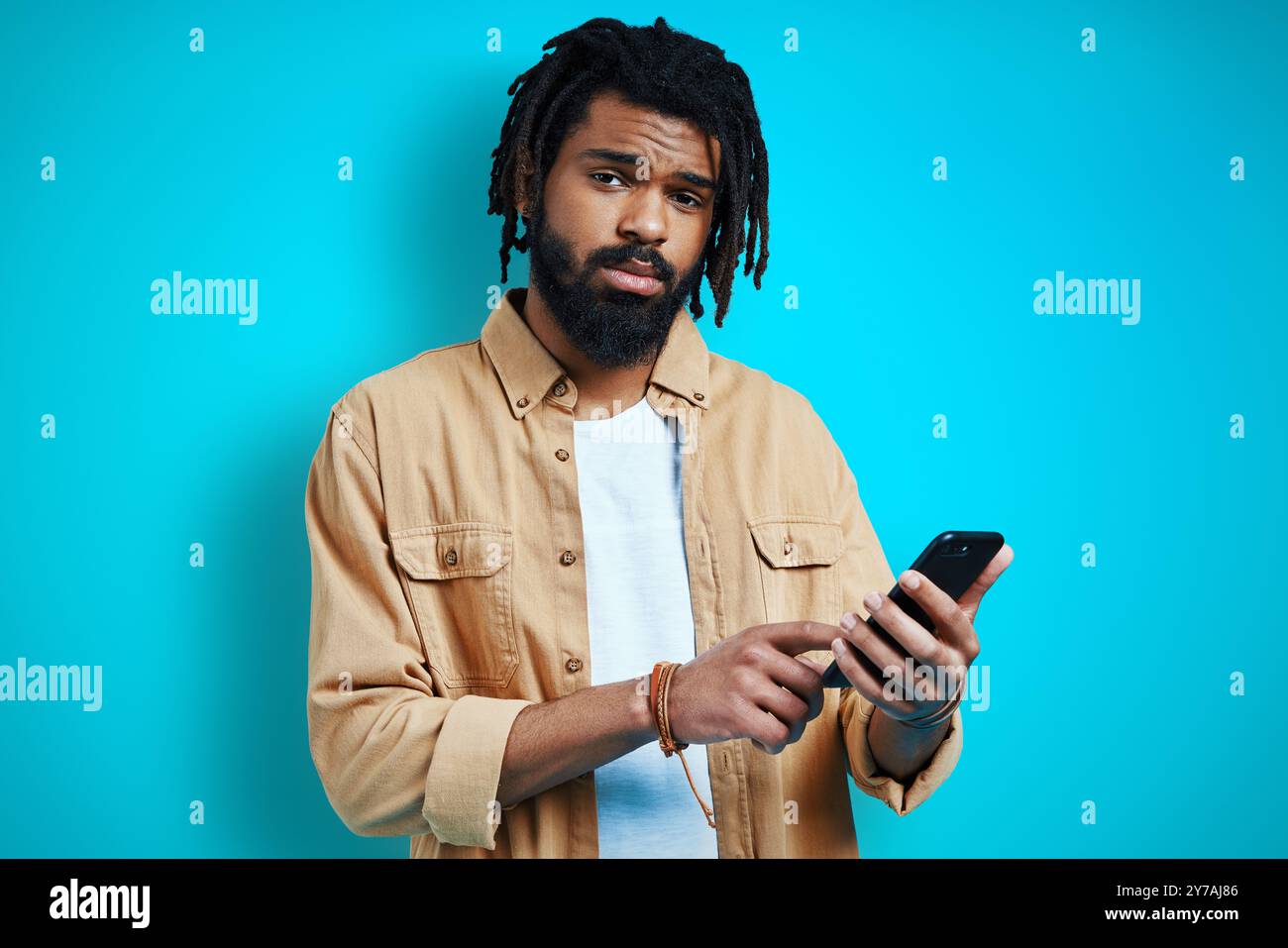 Charming young African man in casual clothing using smart phone and looking at camera while standing against blue background Stock Photo