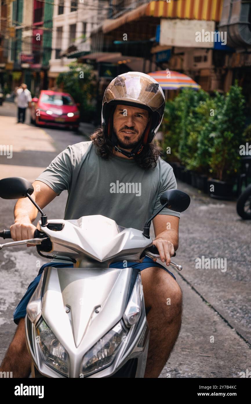 Vertical portrait of mid adult man driving a scooter with an open face helmet in the southeast of Asia; front view in a street of Sukhumvit, Bangkok, Stock Photo