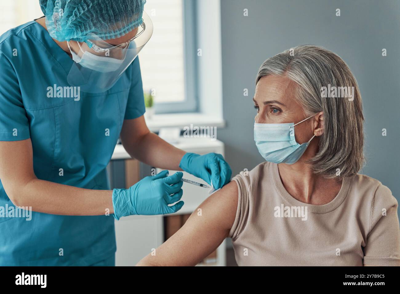 Female nurse in protective workwear making coronavirus vaccination to mature woman while working in the hospital Stock Photo