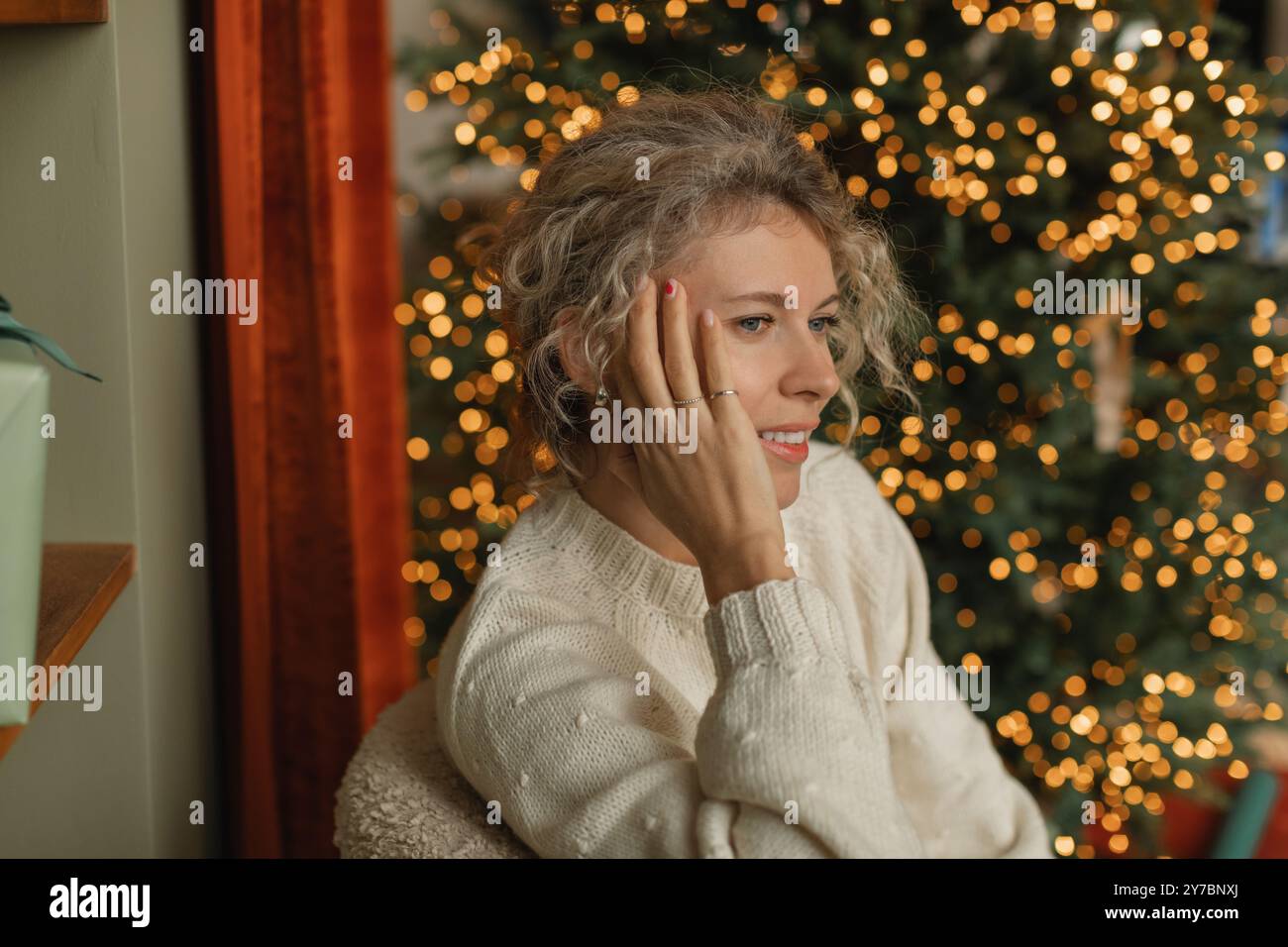 Portrait of a happy woman with her head in her hands sitting on a chair next to an illuminated Christmas tree in a living room Stock Photo