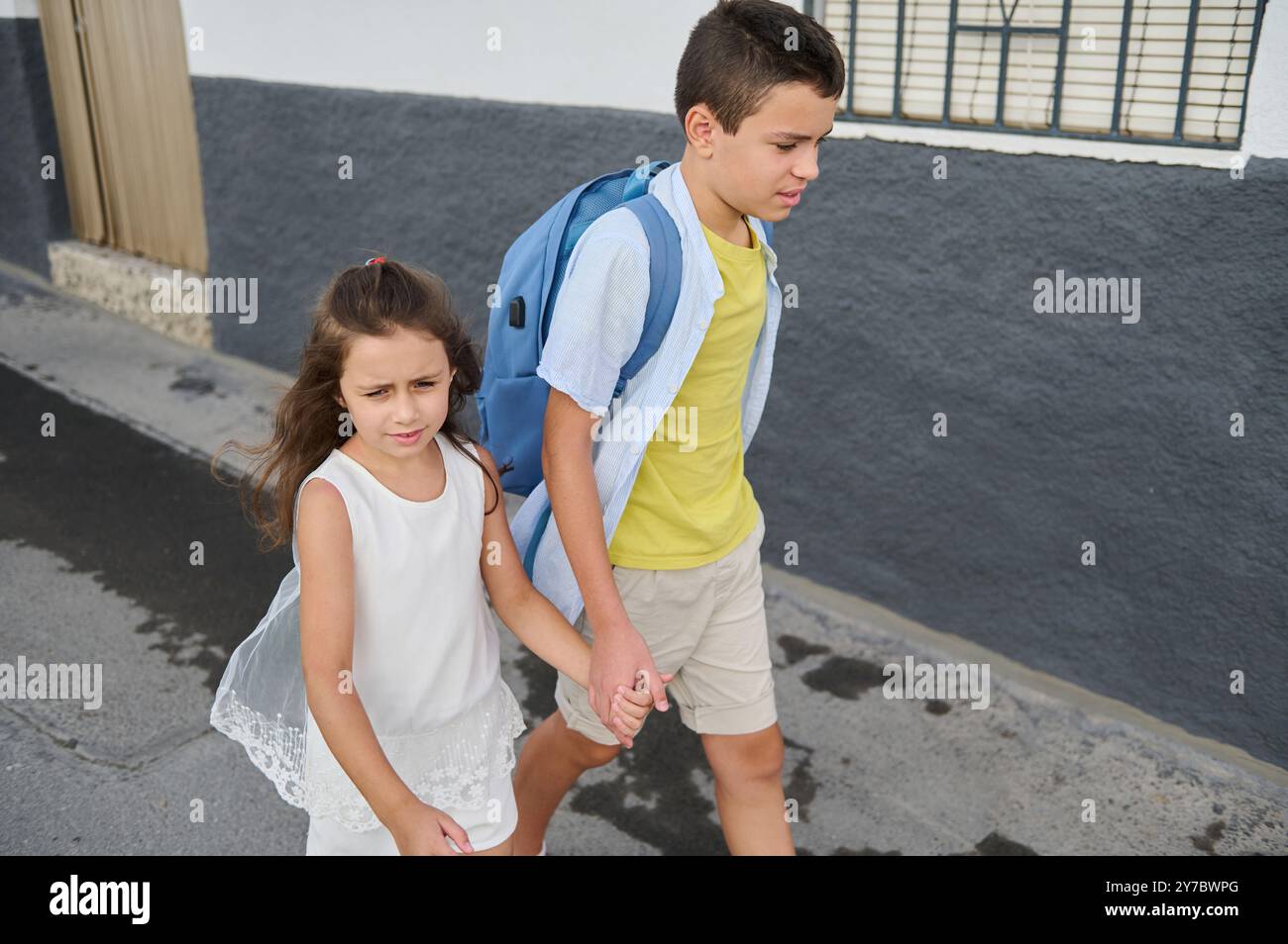 Two siblings walk hand in hand on a city sidewalk, conveying ...