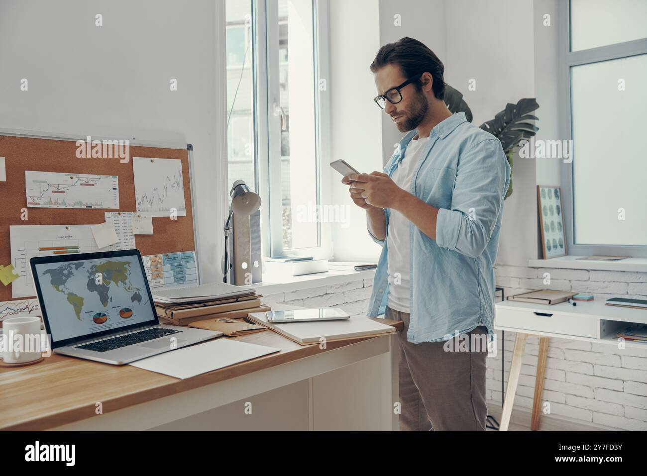 Confident young man using smart phone while standing near his working ...