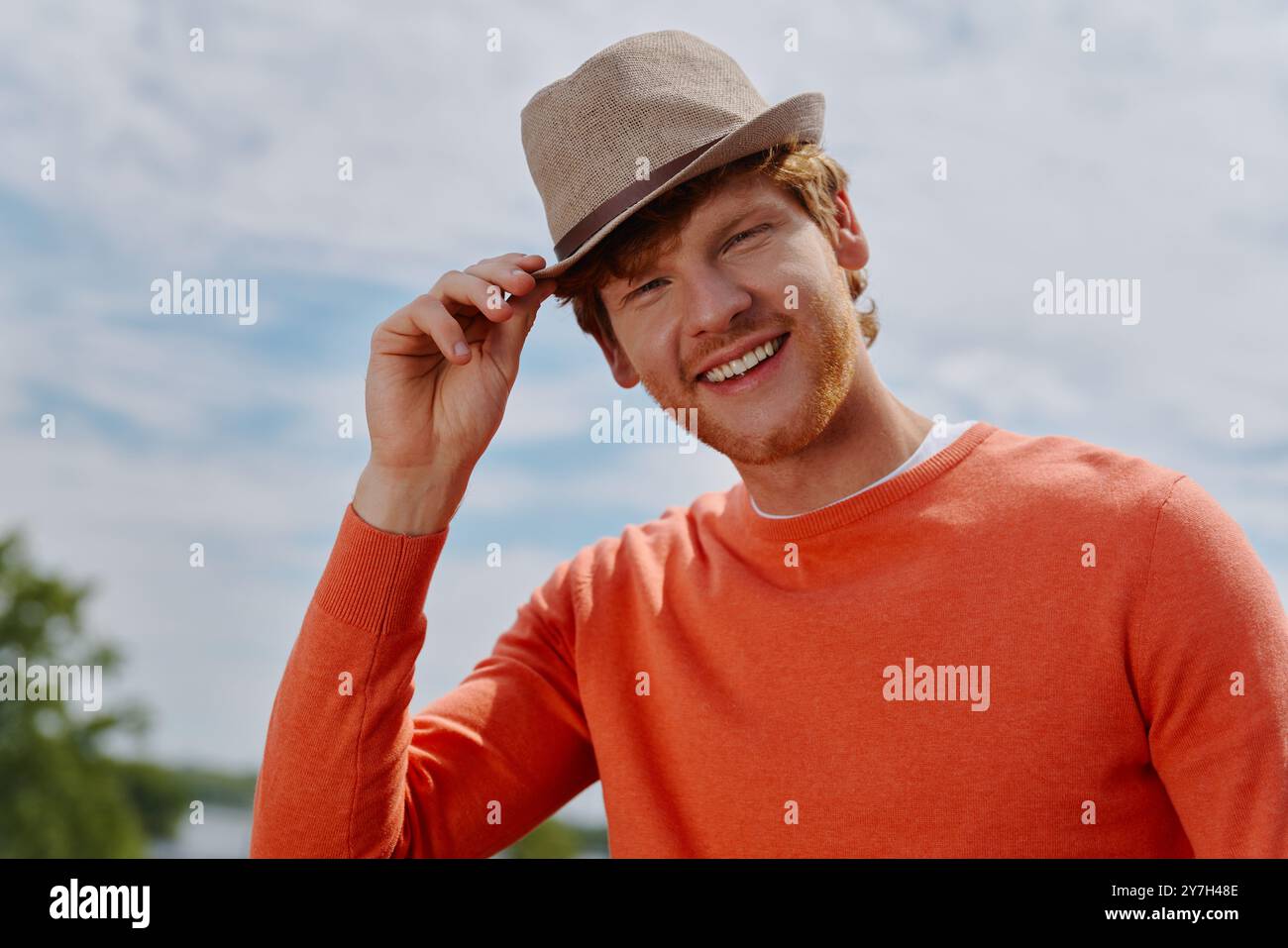 Relaxed young redhead man adjusting his hat and smiling while standing ...