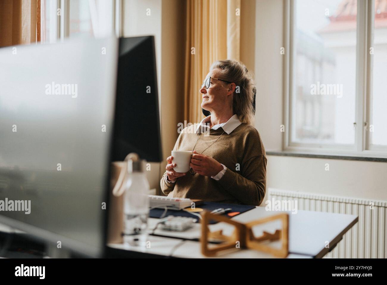 Thoughtful businesswoman holding coffee cup while sitting on chair near desk at office Stock Photo