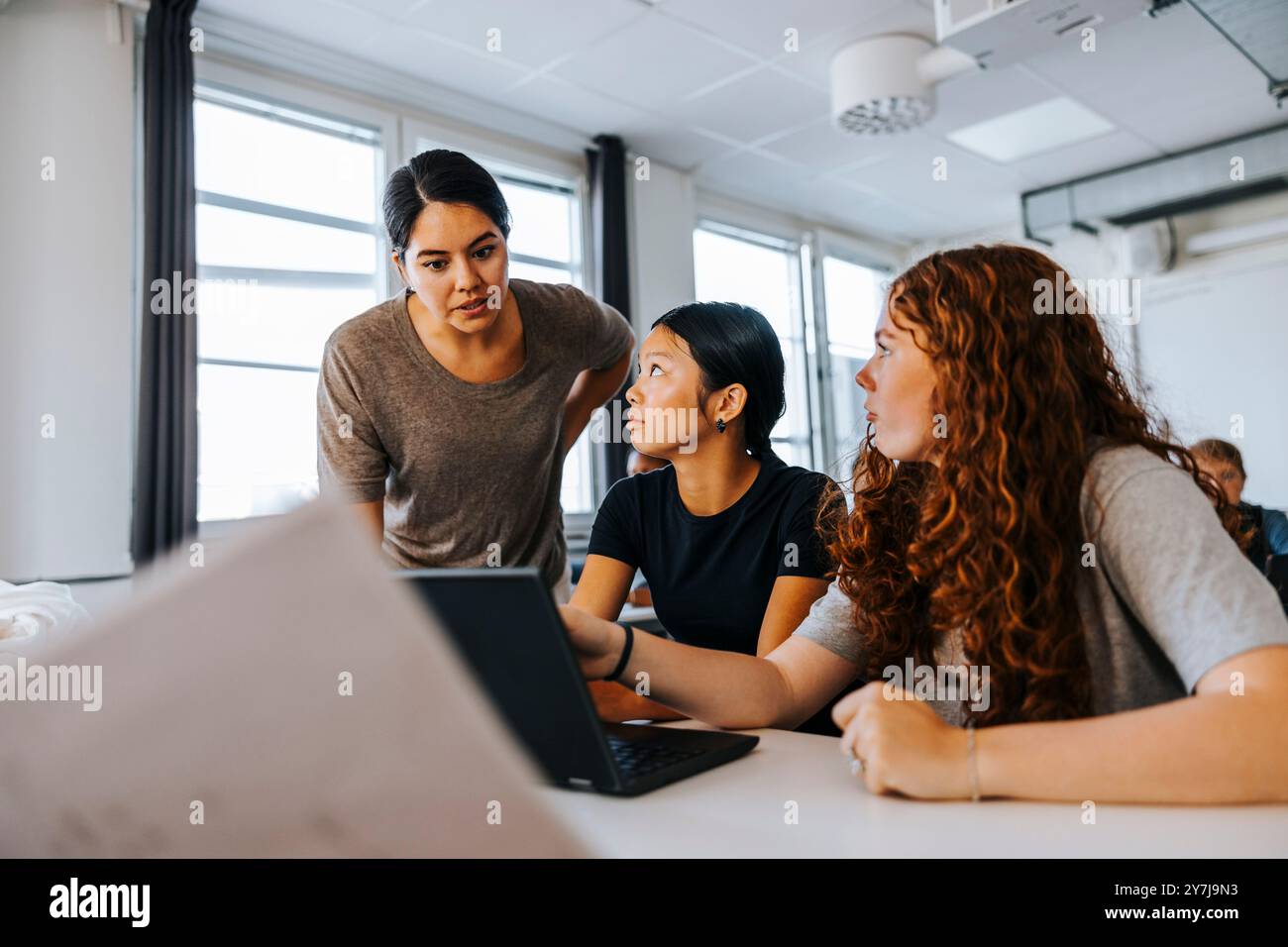 Focused teacher guiding female junior high students on laptop in classroom at school Stock Photo