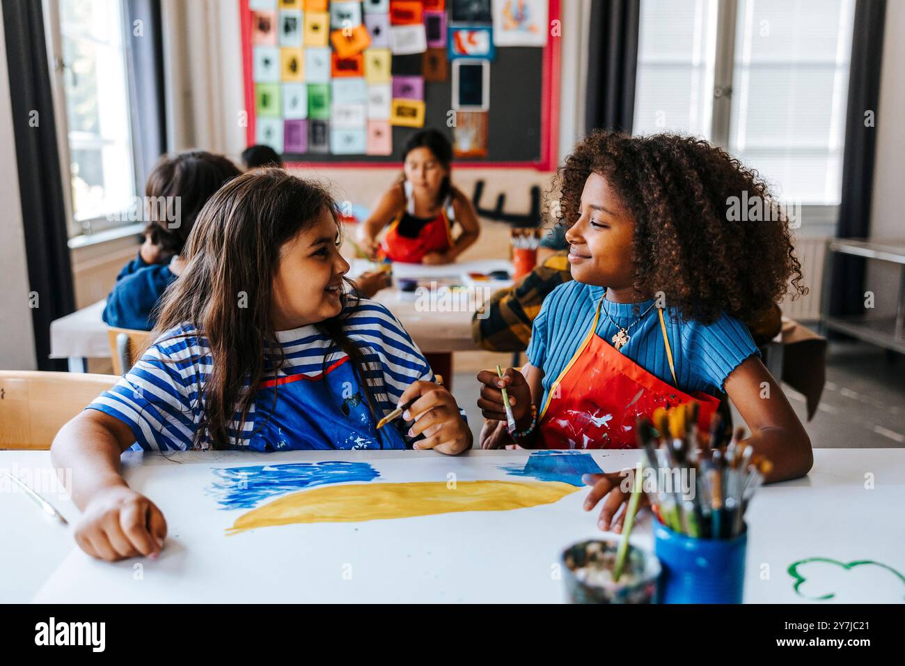 Smiling curly hair girl with female classmate sitting in art class at ...