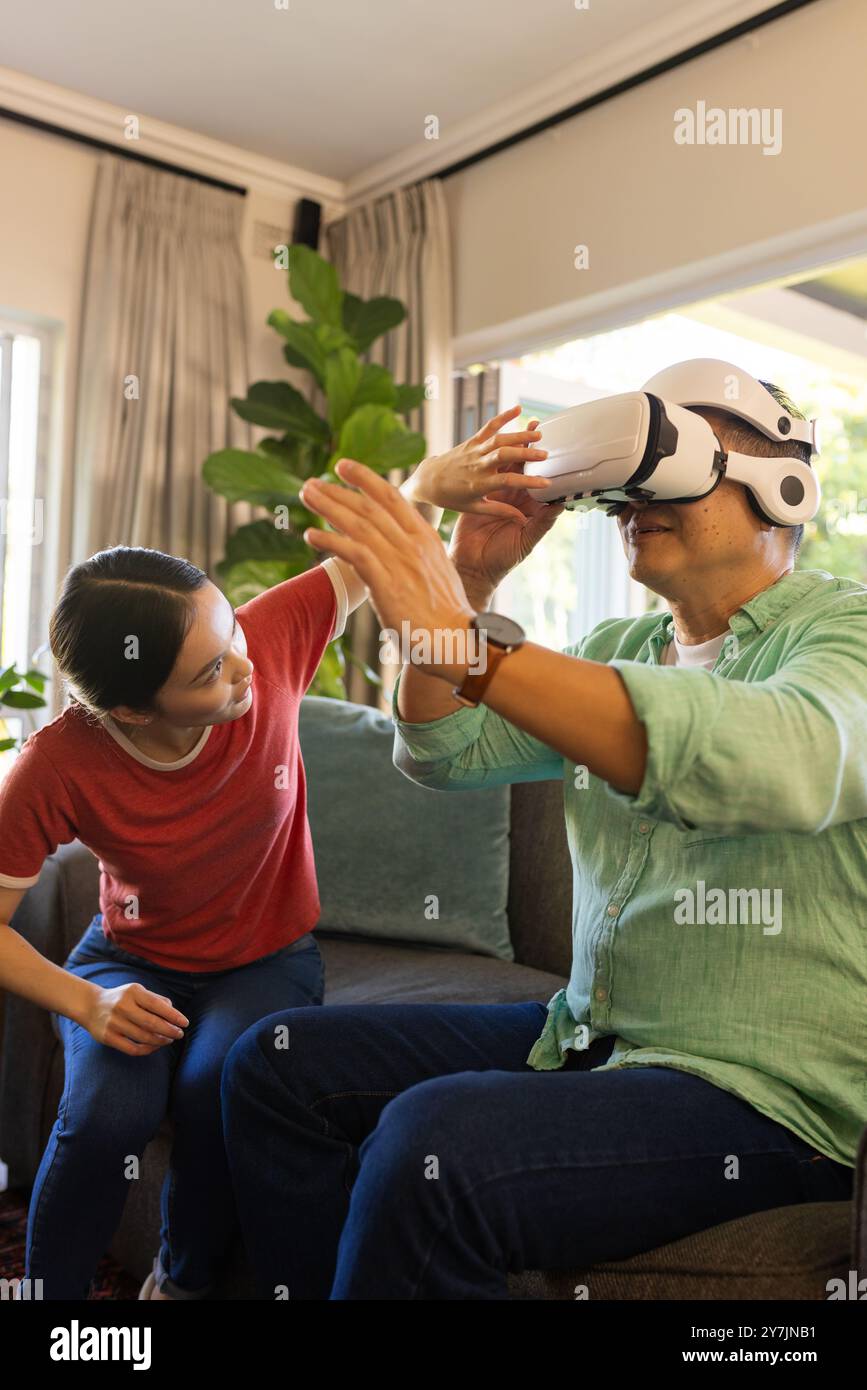Using VR headset, father exploring virtual reality while daughter assists at home Stock Photo