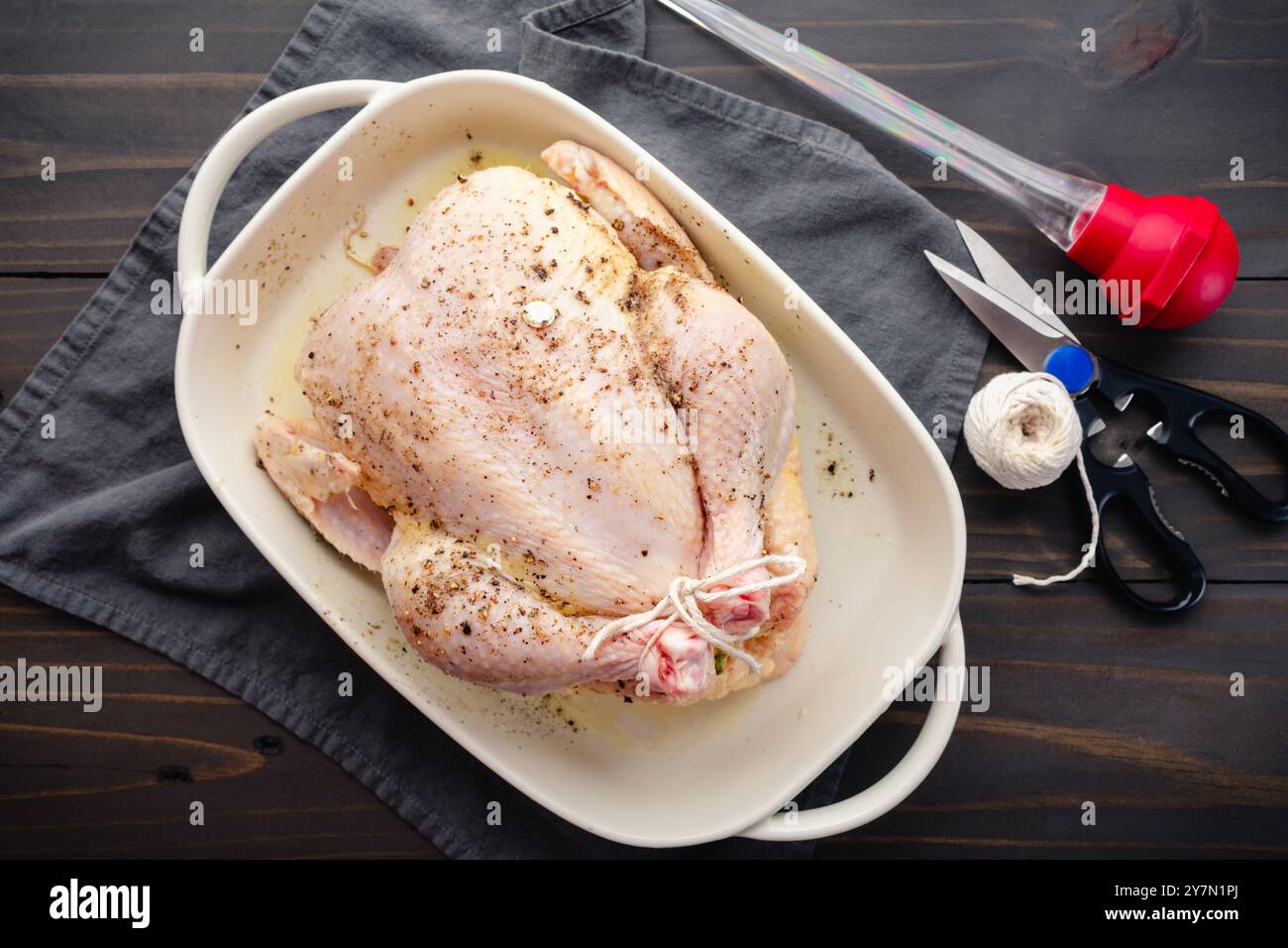 Seasoned Raw Chicken in a White Baking Dish: Whole chicken with tied legs covered in melted butter, sea salt, and black pepper in a baking pan Stock Photo