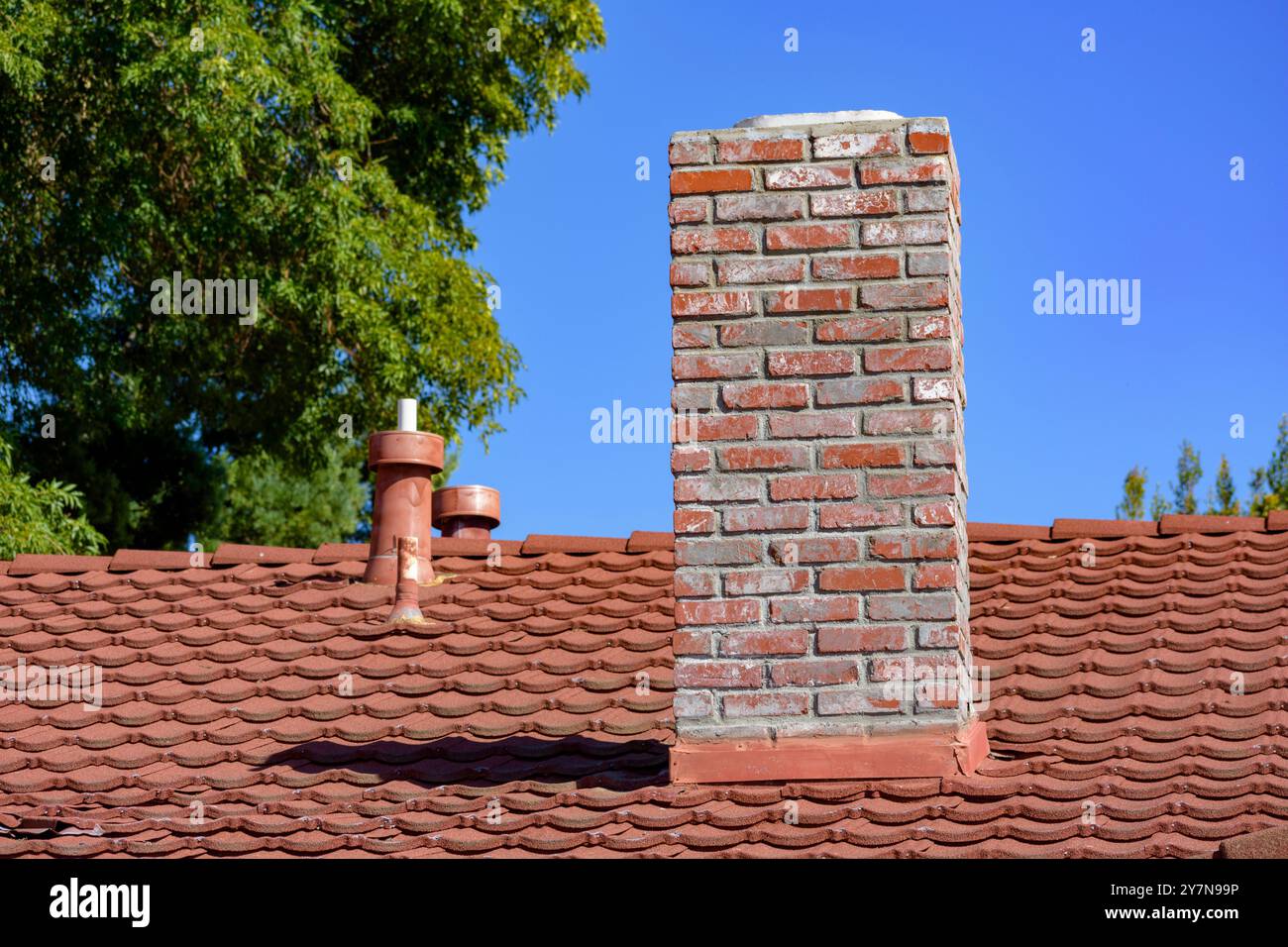 A residential roof with red tiles, a brick chimney and metal vents ...