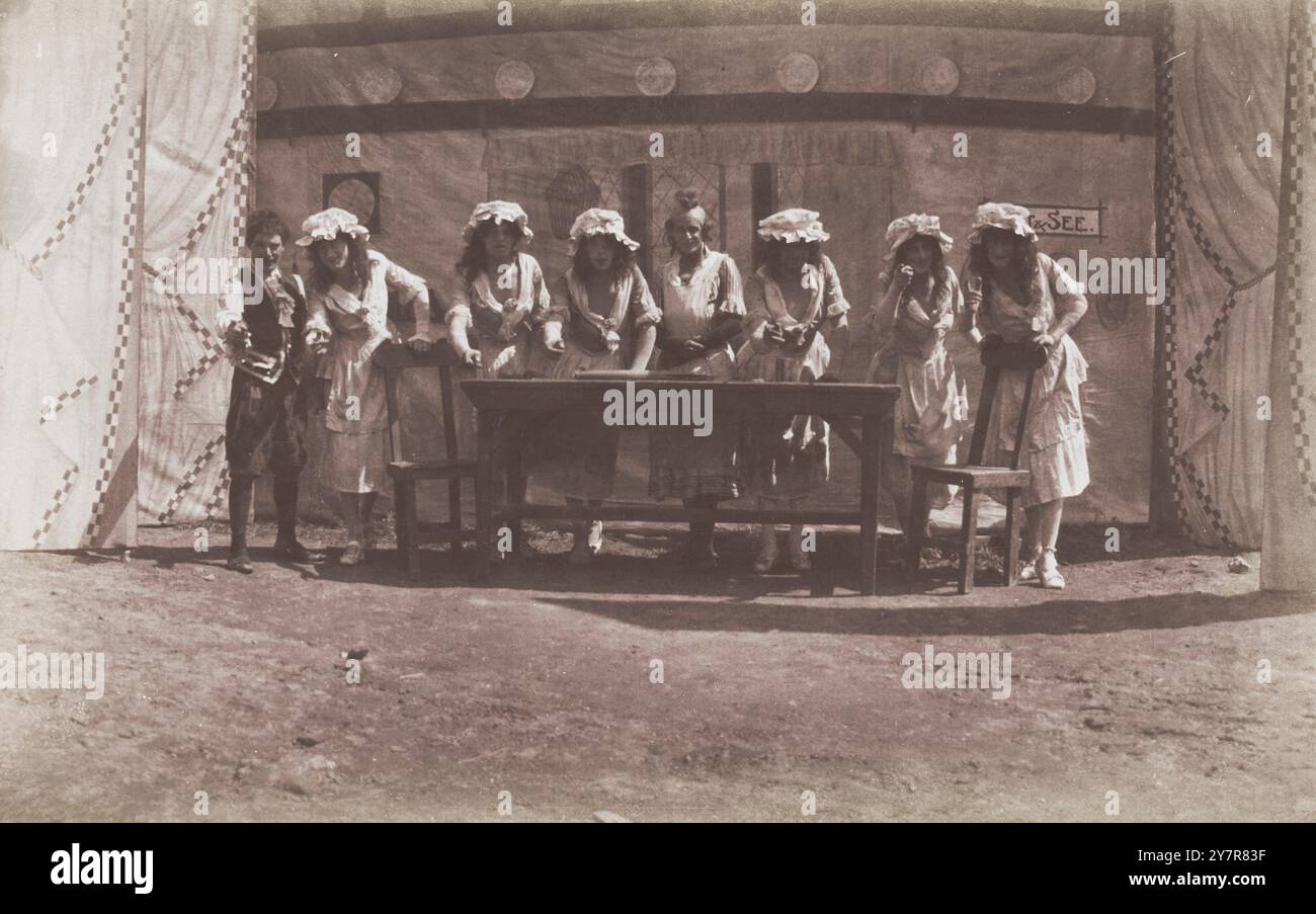 British servicemen in drag acting out a cooking class. Photograph, 1910-1919. Men in drag behind a long table. with a large fabric backdrop. The actors are wearing mob caps and have their sleeves rolled up, representing a 'Cookery class' (according to inscription on verso). This photograph was taken during a British concert party somewhere in the Middle East towards the end of World War I. Stock Photo