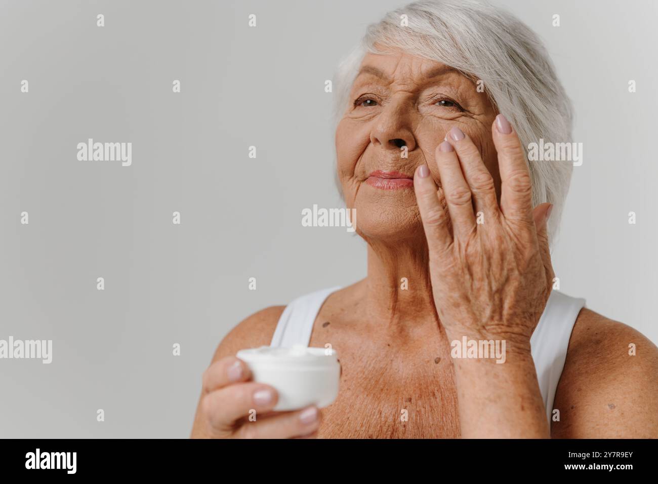 Confident senior woman applying anti-aging cream on face against grey background Stock Photo