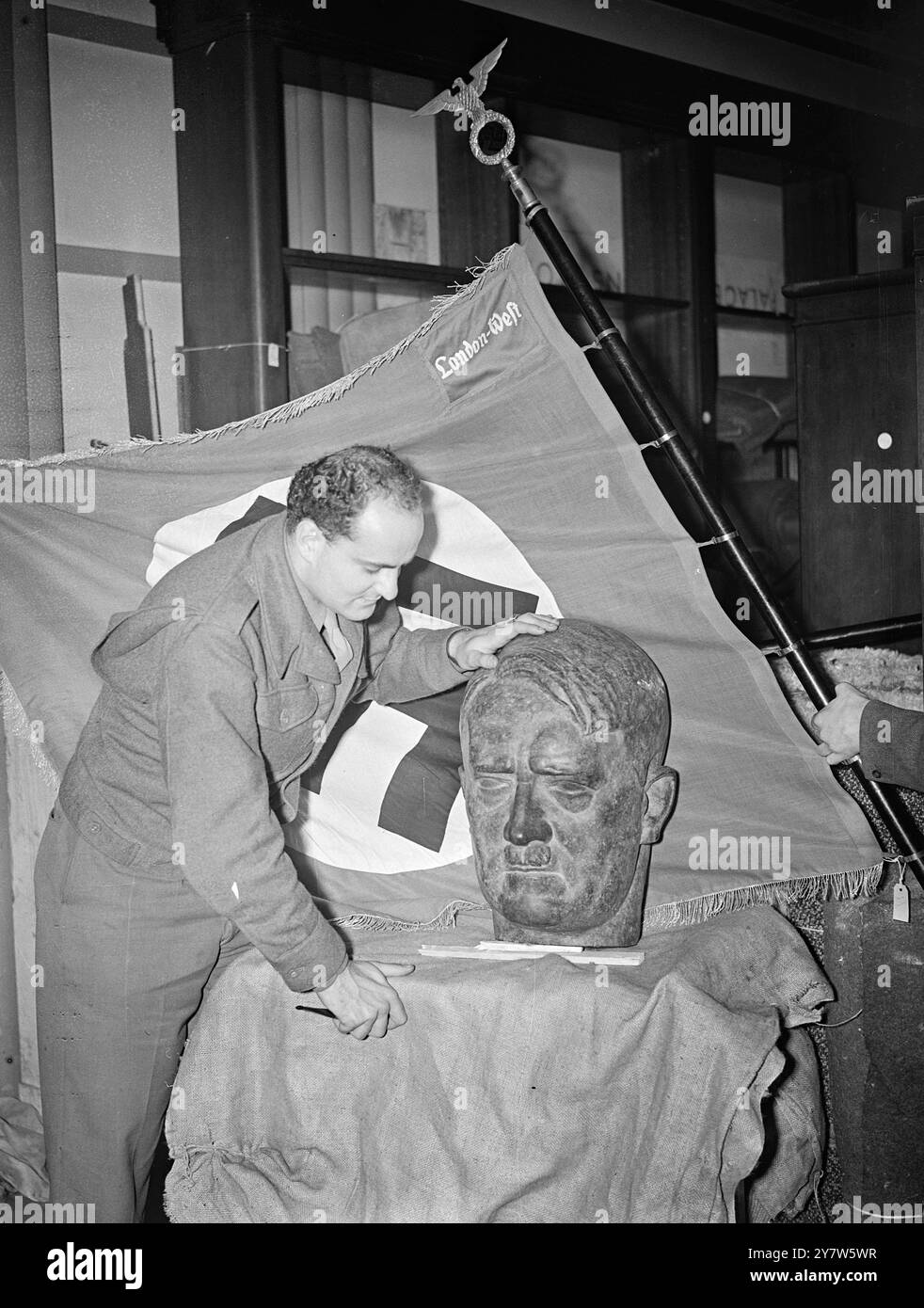 GERMAN EMBASSY FURNISHINGS PREPARED FOR AUCTION Photo Shows: Pfc. Leroy V Anderson of Washington DC, an American solider working with Messrs, Knight Frank and Rutley under the American Army 'on the job' educational scheme, examines a large bronze bust of Hitler among the effects of the former German Embassy in London to be sold by auction at Earl's Court. Behind is a Nazi flag bearing the inscription in German 'West London' - probably intended for use after the 'capture of London'. Photographs taken at the Brasserie, Earl's Court, London, where the sale will be held and the auction broadcast t Stock Photo