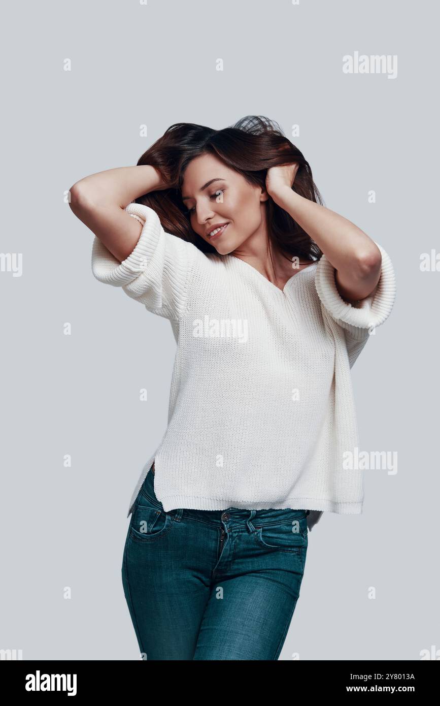 Simply stunning. Attractive young woman keeping hands in hair and smiling while standing against grey background Stock Photo