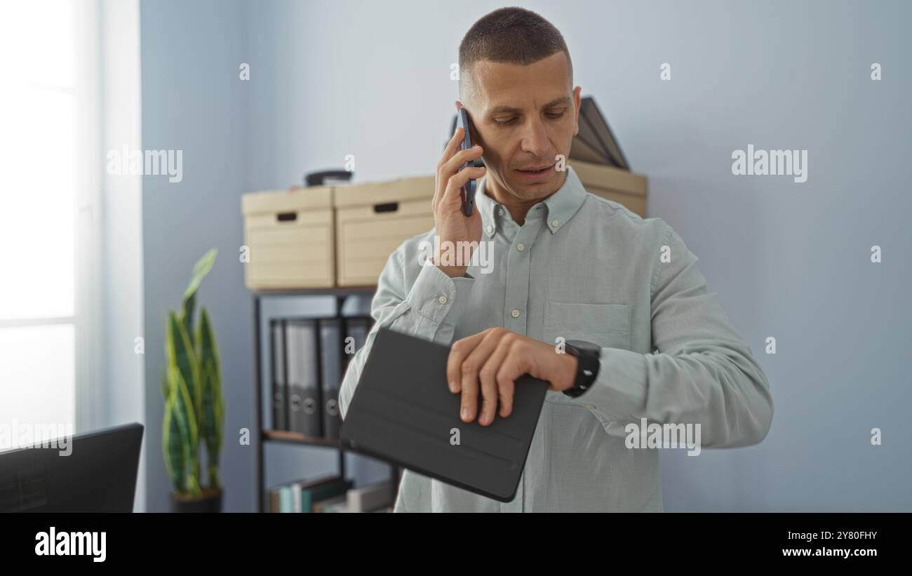 Young man in office checking time on smartwatch while talking on phone, with office furniture and plant in background Stock Photo