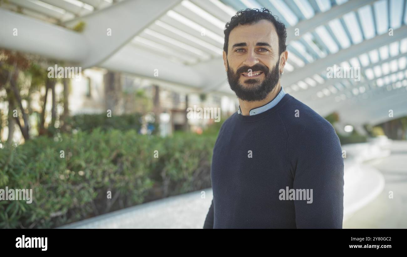 Smiling bearded man in casual attire enjoys a sunny day at a modern urban park. Stock Photo