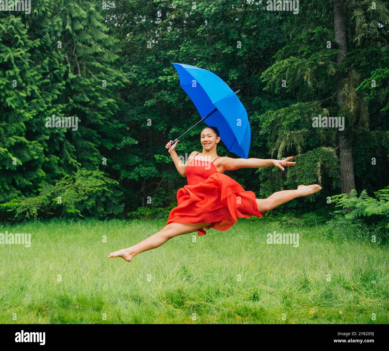 Woman in a red dress joyfully leaping with a blue umbrella in a lush green park. Stock Photo