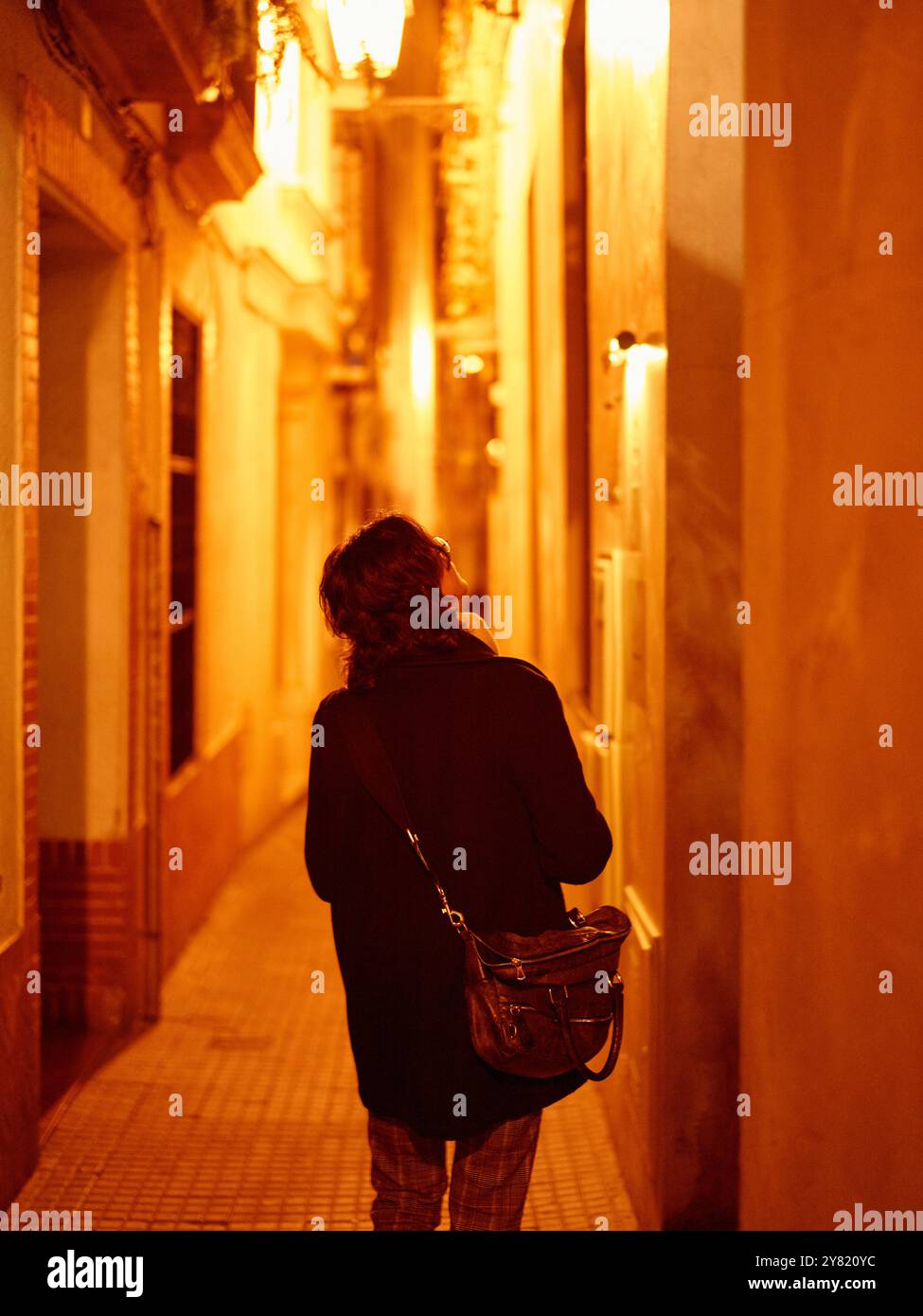 Person stands in a narrow alleyway at night, illuminated by a warm glow from the surrounding lights. Stock Photo