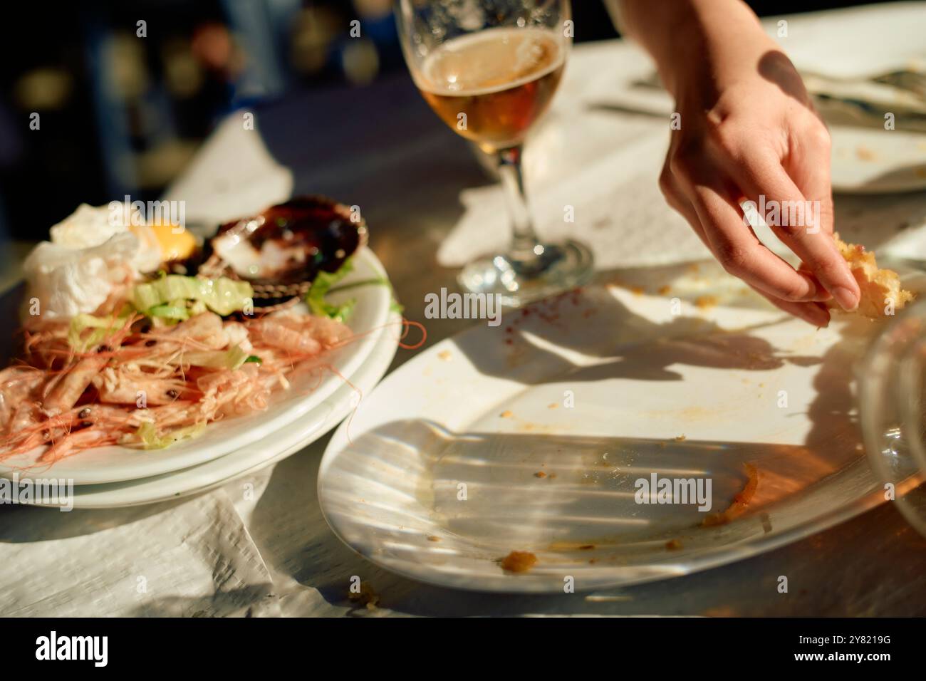 Close-up of a dining table with remnants of a seafood meal, a hand grabbing food, and a glass of beer. Stock Photo