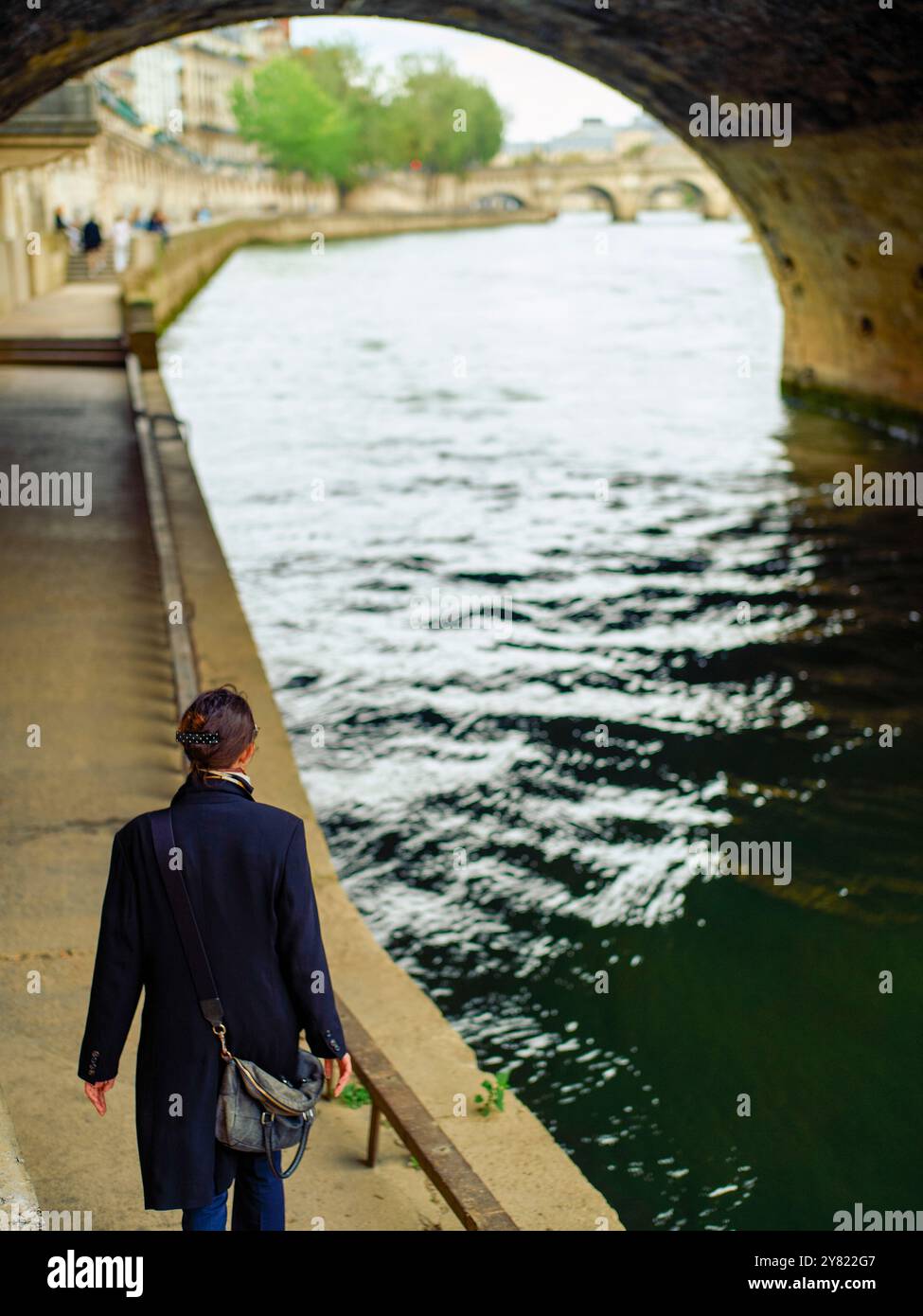 A person walks along a riverside pathway under an arched stone bridge ...
