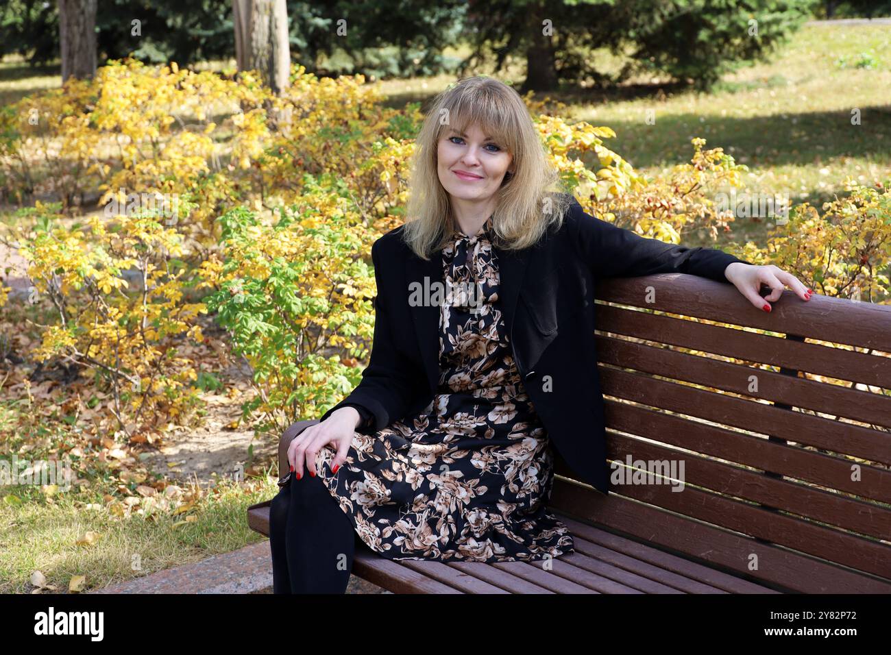 Outdoor portrait of smiling caucasian attractive girl with blond hair sitting on a bench in autumn park Stock Photo