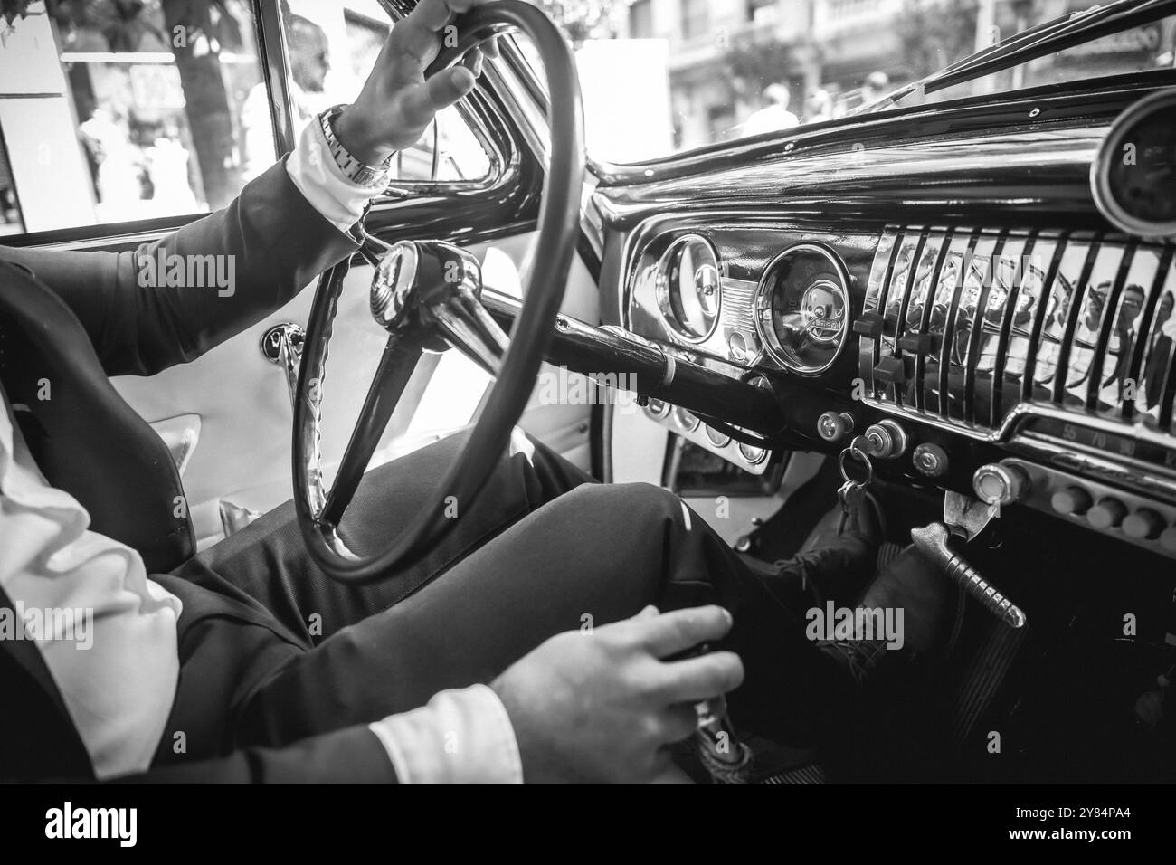 A man in a suit is driving a vintage car. The car is black and white. The man is wearing a watch Stock Photo