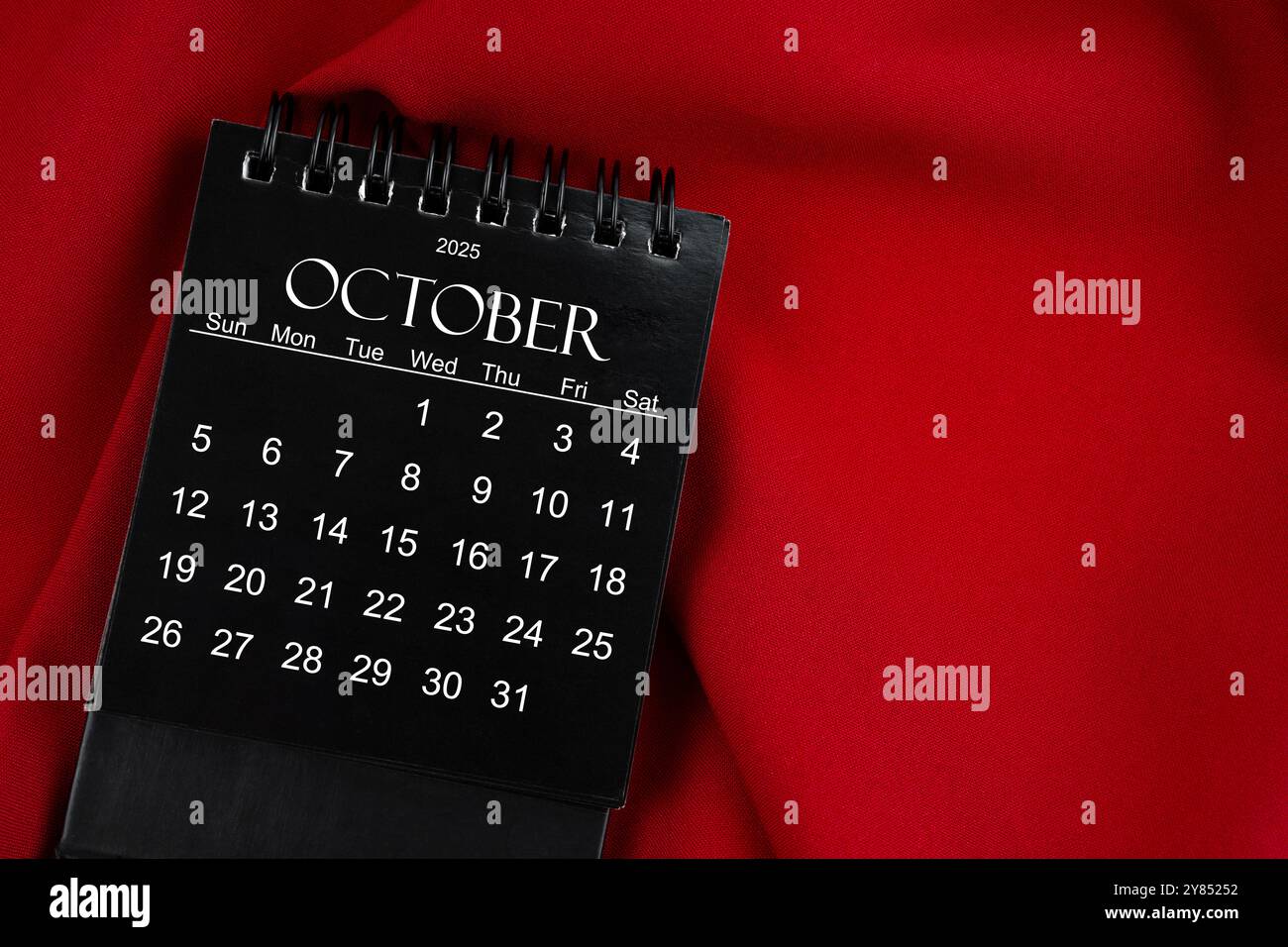 October 2025 Black spiral desktop calendar on red textile background