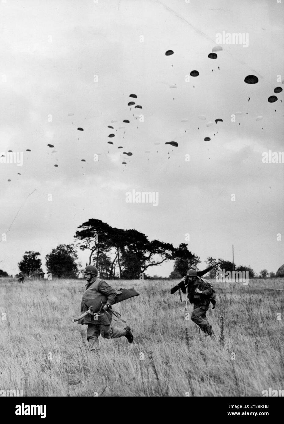 'Part Time' Parachutists in Arnhen Day Exercise -- Two recently landed parachutists running to take up their positions while above them hundred more men float down, The T.A. Brigade actually doing this drop over the Stanford (Norfolk) training area is the 44 the (comprising  two London and one Hampshire Battalions) part of Britain's only Airborne Division T.A.- the 16th. 1,200 Territorials yesterday parachuted down upon their 'enemy' in Norfolk, during exercise 'King's Joker' the biggest peacetime T.A. airborne exercise ever held. The man and their equipment dropped from a height of 800 feet f Stock Photo