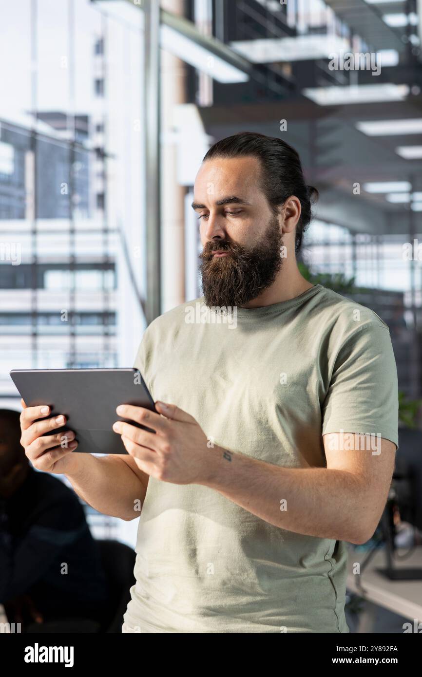 Man working in office developing software in Java programming language using device. Programmer walking through workspace with tablet, thinking how to solve coding problems Stock Photo