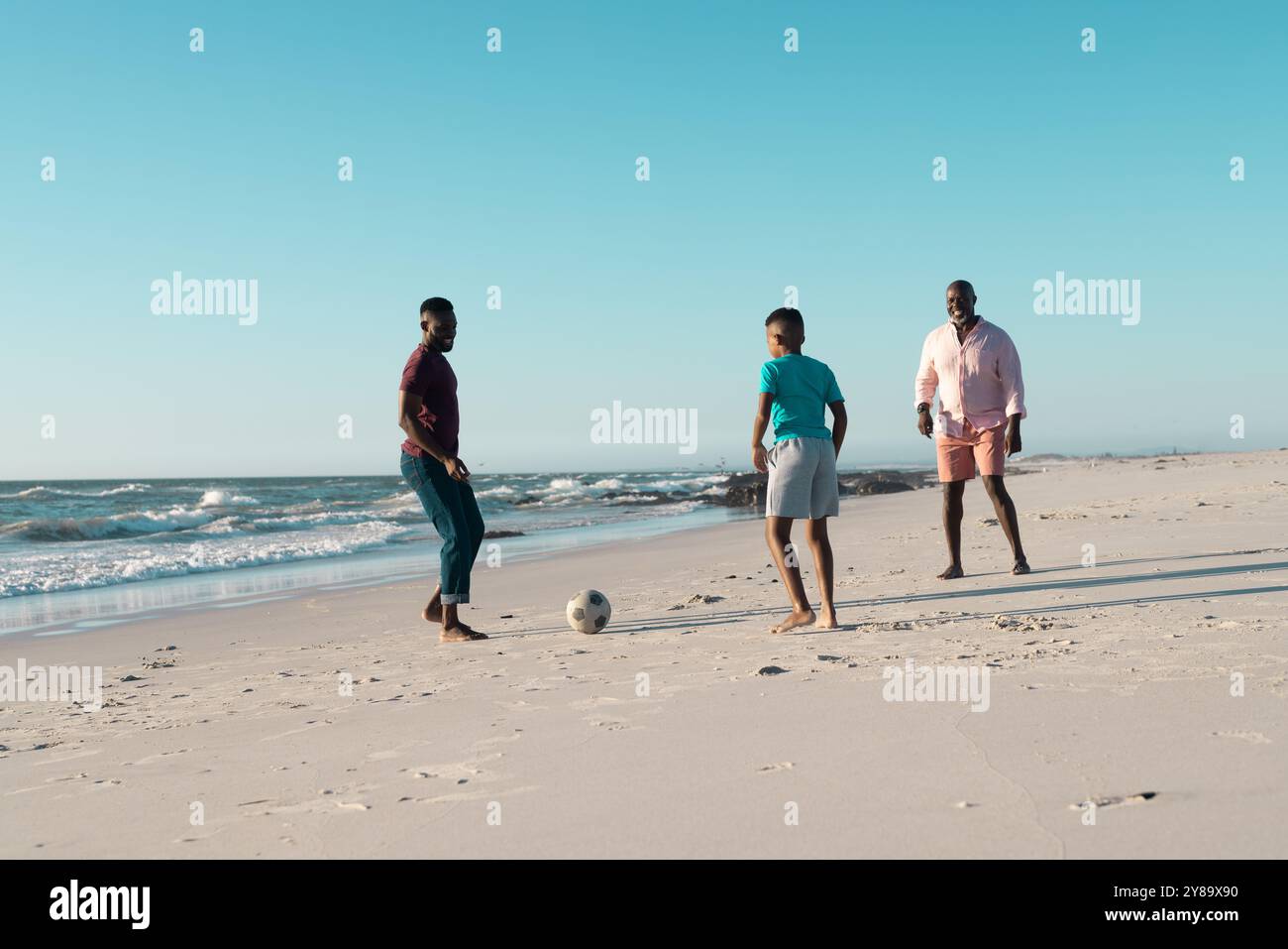 African american boy playing soccer with father and grandfather at beach against clear sky Stock Photo