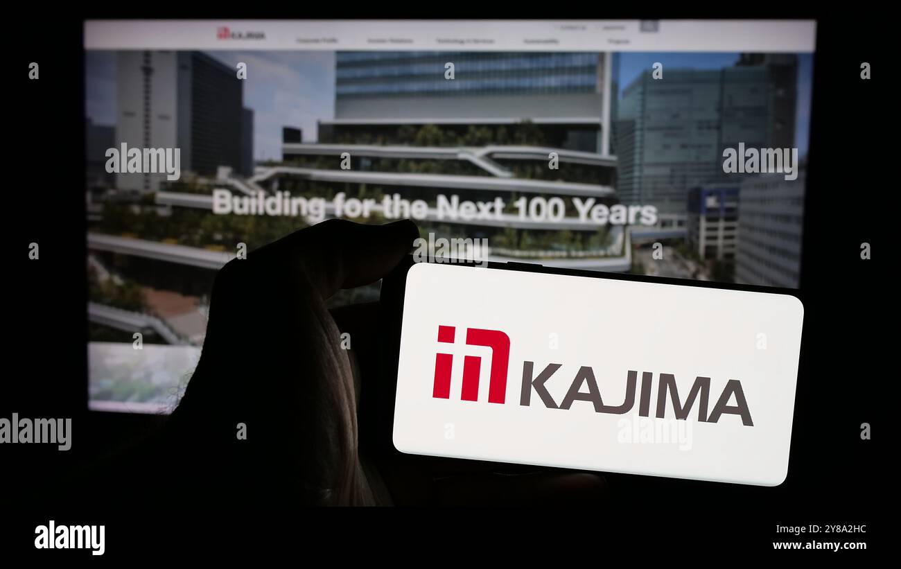 Person holding mobile phone with logo of Japanese construction company Kajima Corporation on screen in front of web page. Focus on phone display. Stock Photo