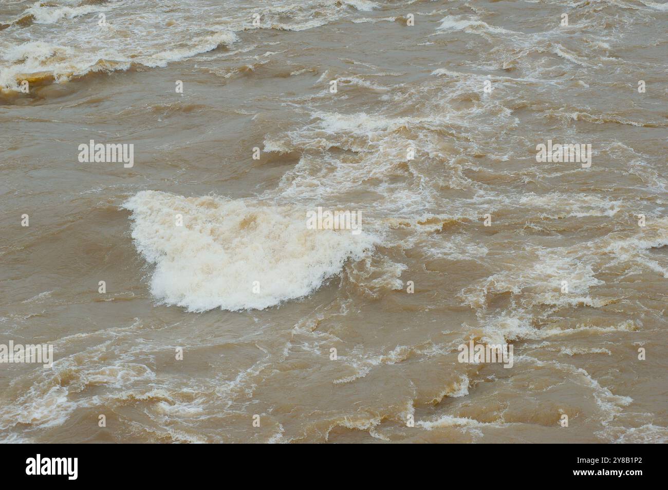 Medium overhead view looking down at a swift fast moving, churning muddy clay brown river water flow rolling with White caps. Stock Photo