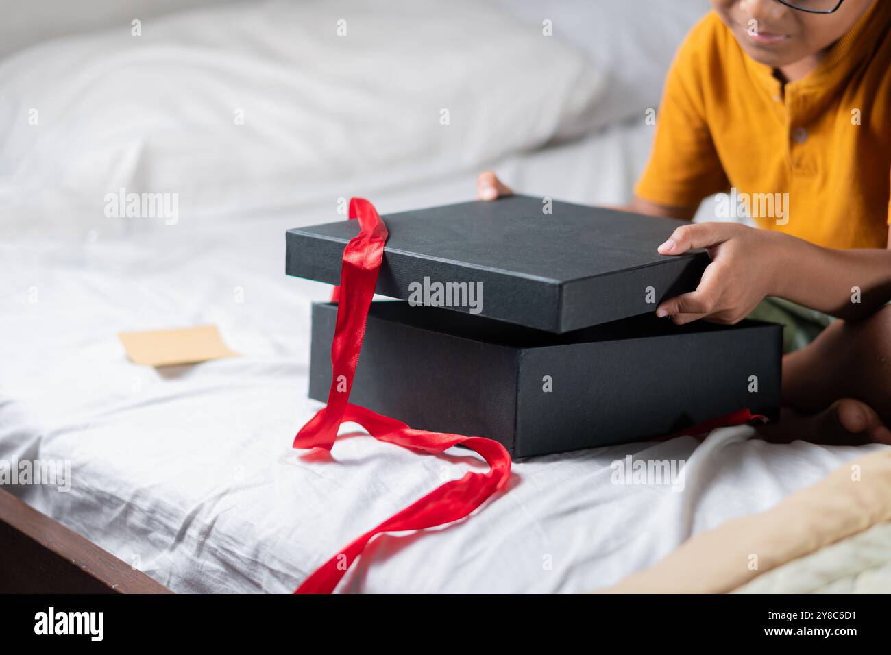 A child is sitting on a bed, excitedly opening a black gift box with a red ribbon. The child is wearing an orange shirt and glasses, showing a joyful Stock Photo