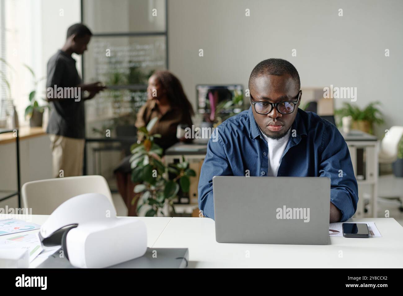 Young African American man with glasses and dressed in casual clothes working via silver laptop while his colleague chatting in blurred background Stock Photo