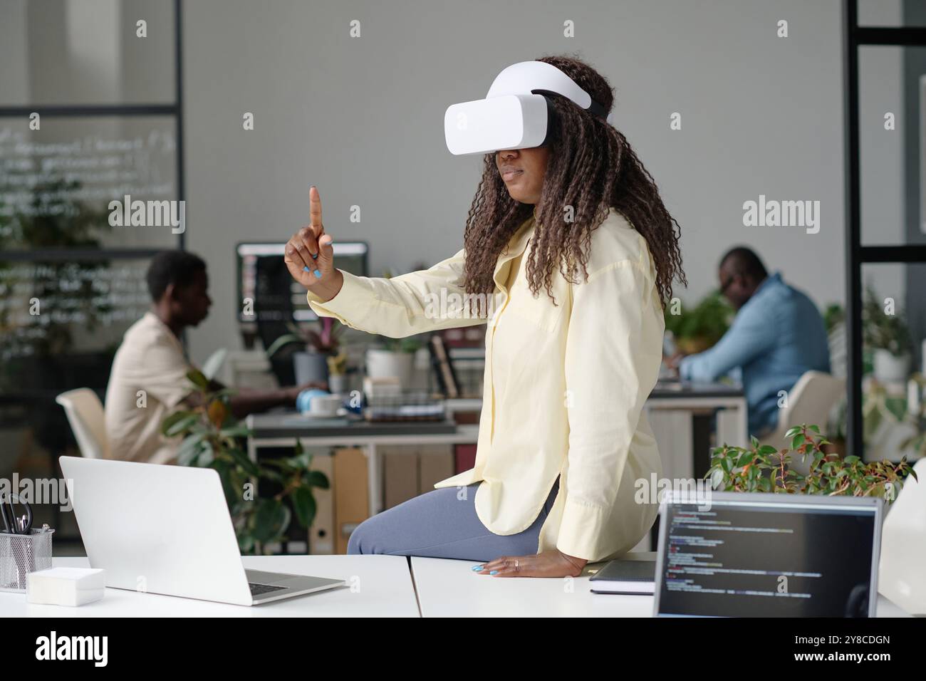Curly haired woman playing video game while sitting on work table with laptops Stock Photo