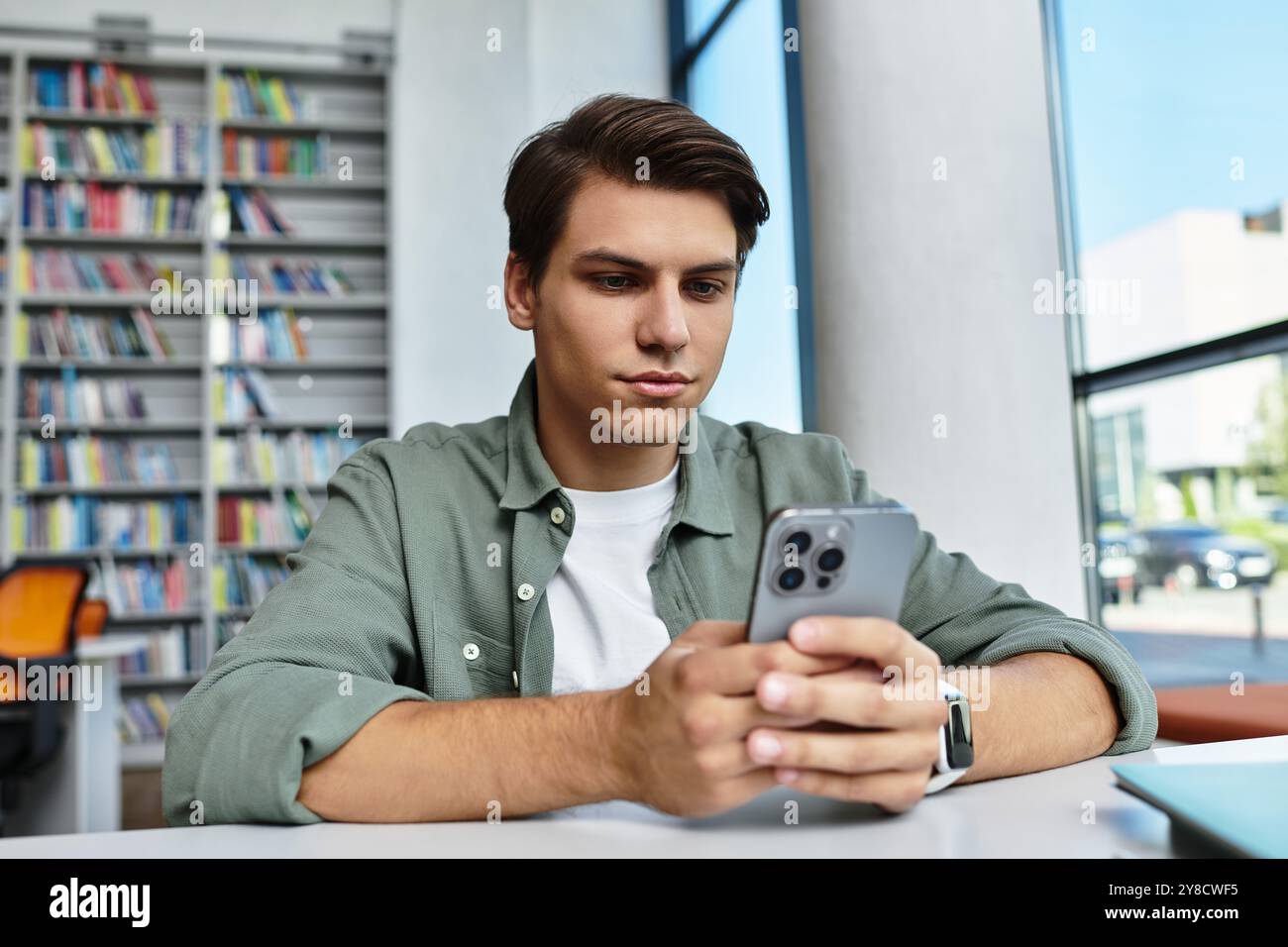 A focused student studies intently while using his smartphone in a vibrant library setting. Stock Photo