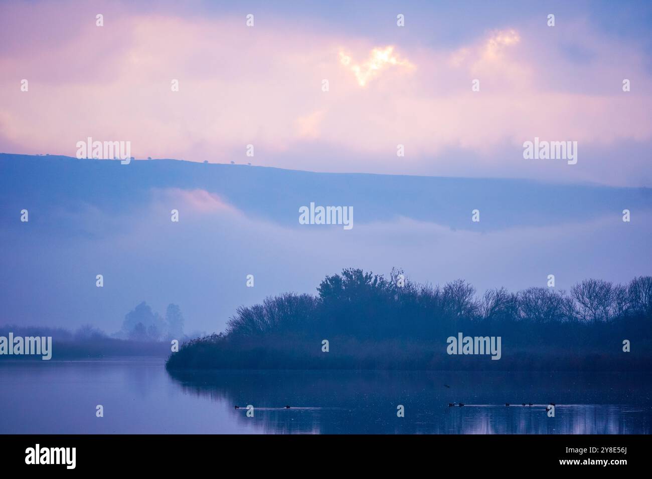 A tranquil scene of a mist-covered lake with hills in the background, captured at sunrise. The soft pastel colors of the sky reflect on the calm water Stock Photo