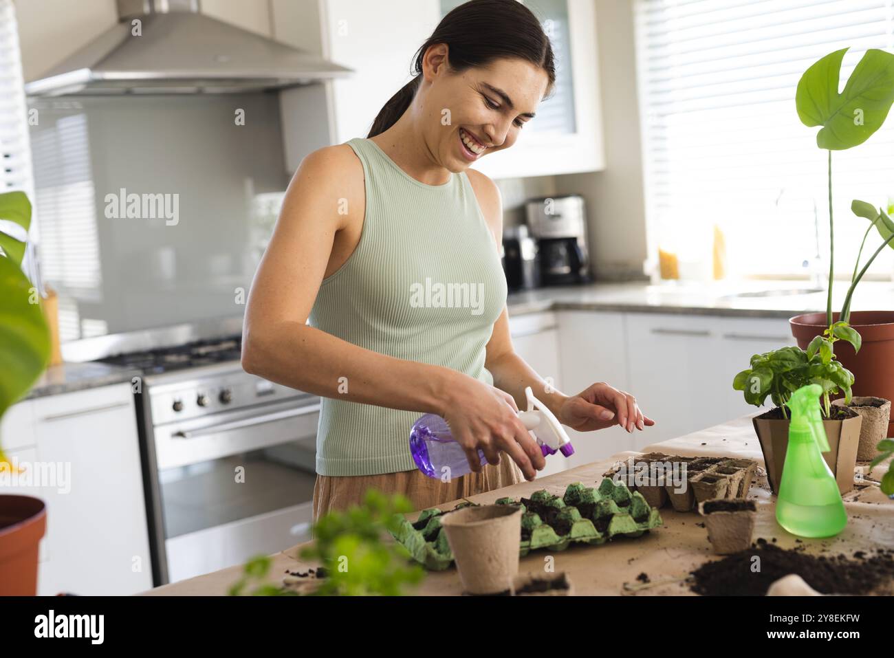 Cheerful caucasian young woman spraying water in seedling tray on table while planting at home Stock Photo