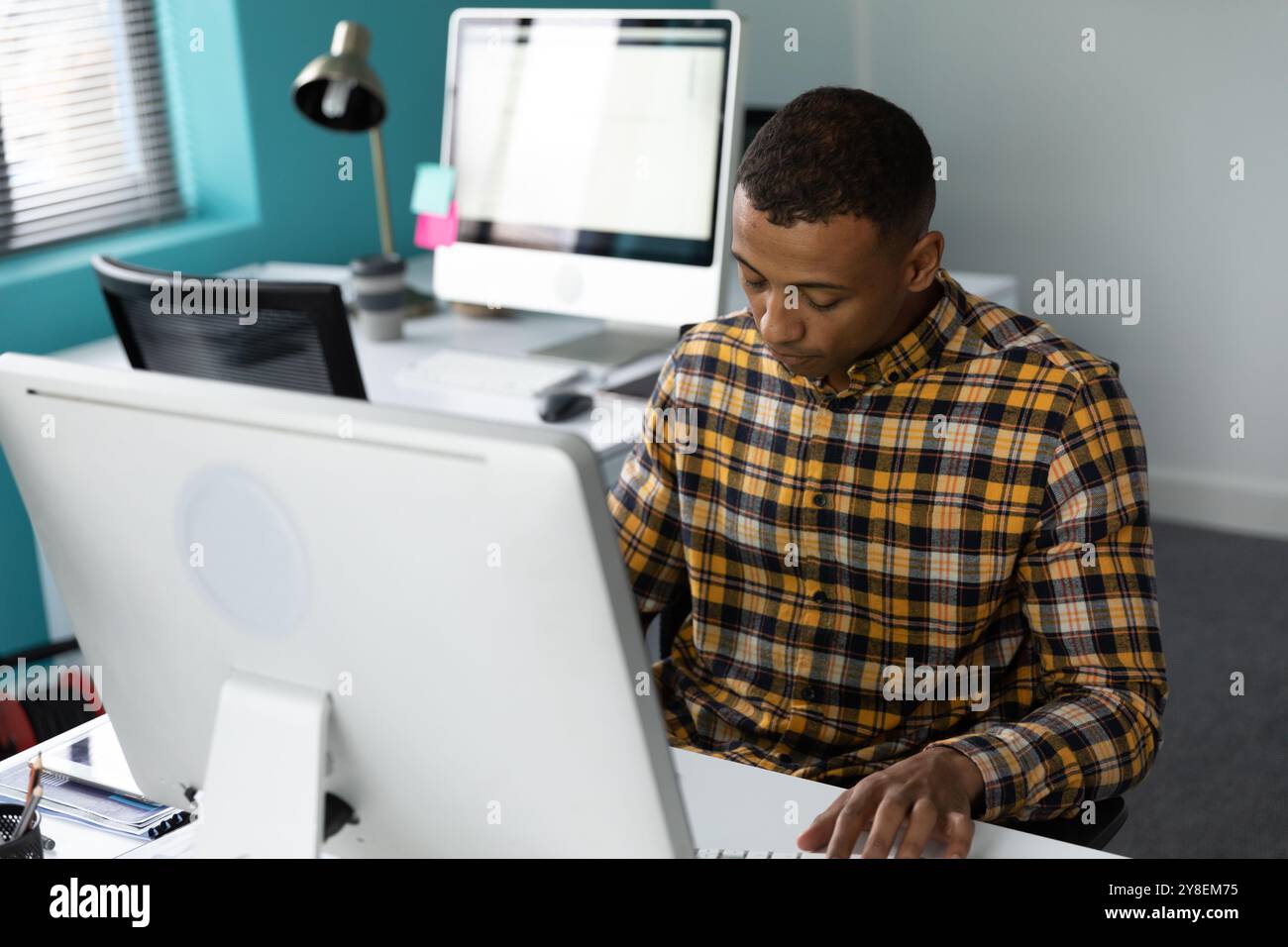 Front view of a young African American man sitting at a desk using a computer in the modern office of a creative business, with an empty workstation i Stock Photo