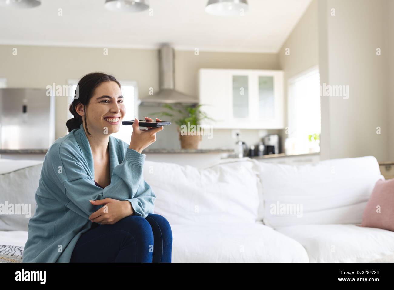 Smiling caucasian young woman talking on speaker over mobile phone while sitting on sofa at home Stock Photo