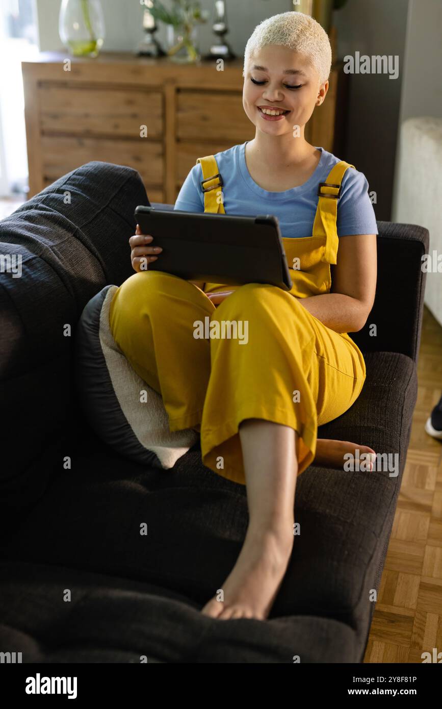 Relaxed fashionable unaltered biracial woman sitting on couch at home using tablet and smiling Stock Photo