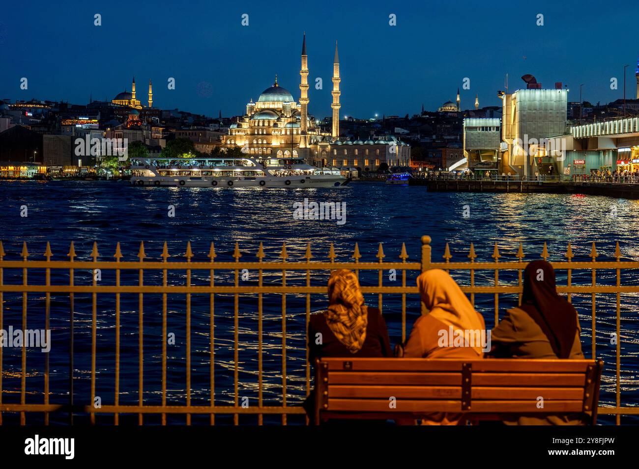 Istanbul. Türkiye. Three Muslim women sitting on a bench in front of the Bosphorus at dusk with the Yeni Cami Mosque on background Stock Photo