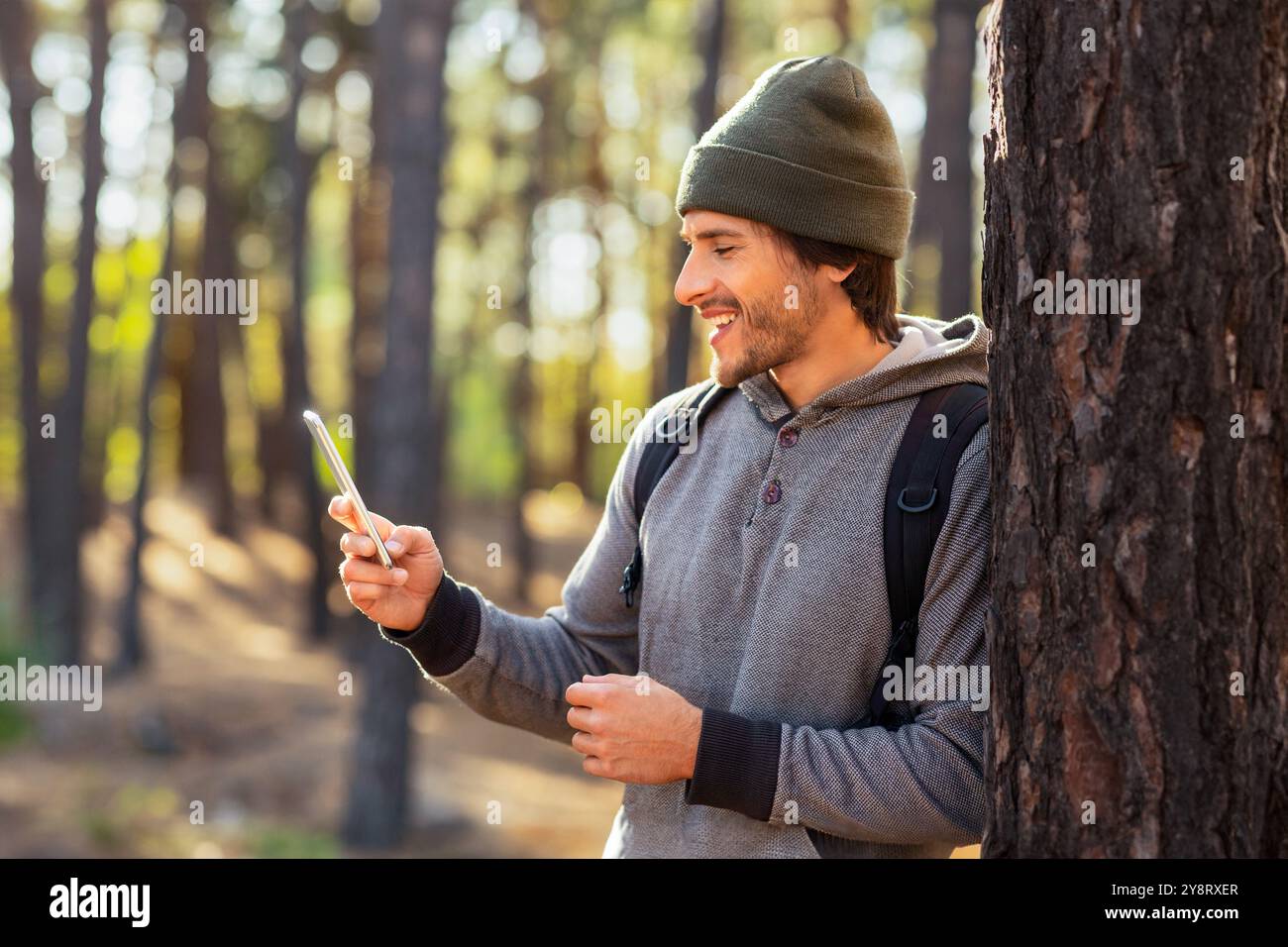 Positive guy leaning on tree in forest, using mobile phone Stock Photo