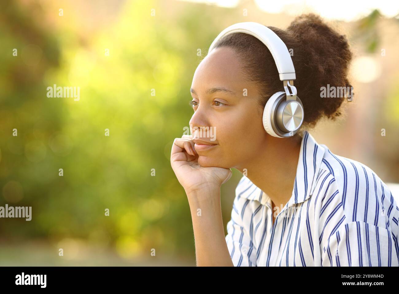 Relaxed black woman sitting in a park wearing headphone looks away ...