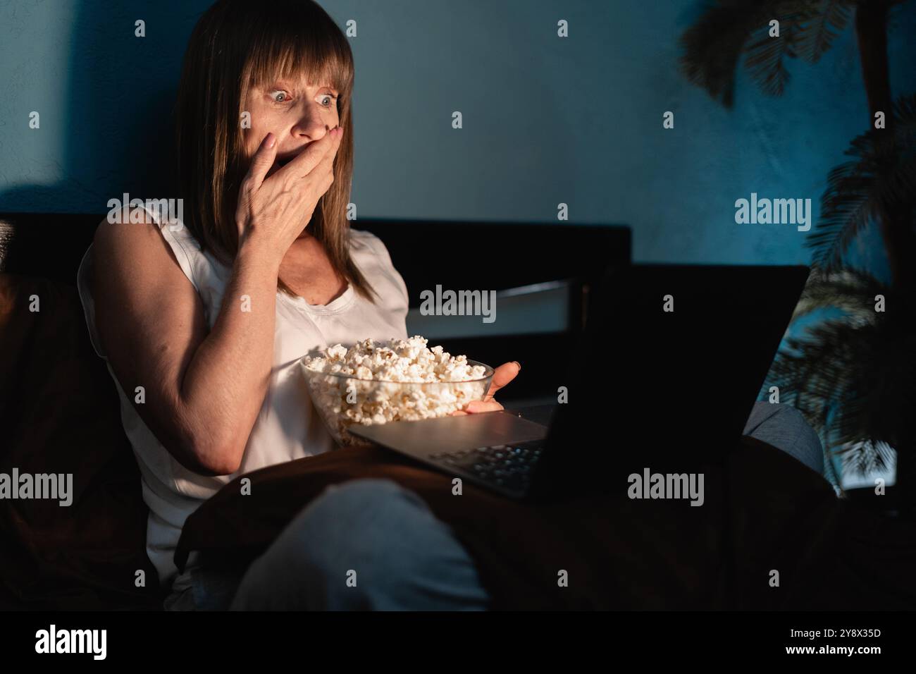 Viewer watching horror series on streaming service, enjoying time in bed. Astonished woman covering mouth in disbelief, shocked by film scene looking Stock Photo