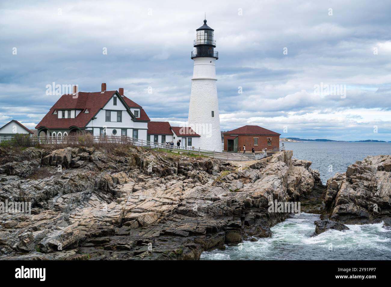 The Portland Head Light stands tall on a rocky coastline overlooking the ocean. People walk along the path near the lighthouse and keeper's house. Stock Photo