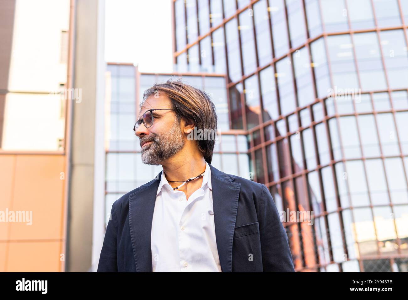 A mature businessman in a stylish black suit looks away thoughtfully, set against a backdrop of modern office buildings. Stock Photo