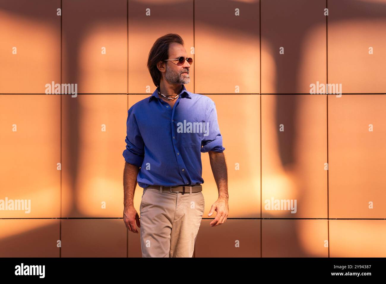 A mature man, dressed in a business casual blue shirt and beige trousers, confidently strolls in a city. He is wearing sunglasses and a modern watch, Stock Photo